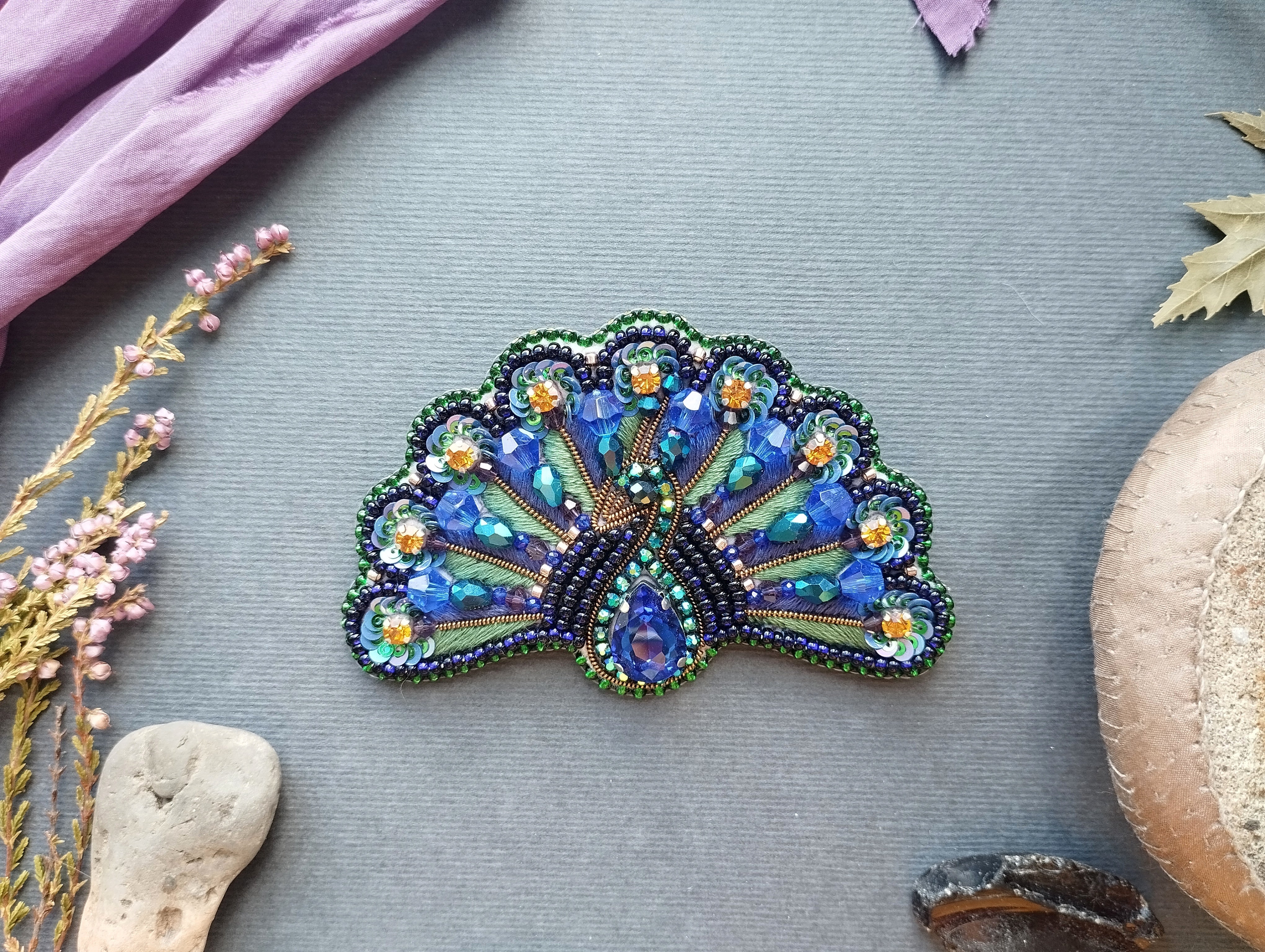 a decorative peacock feather-shaped brooch with intricate beading and gemstone details, placed on a gray background alongside various natural elements such as leaves, flowers, and rocks.